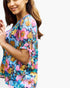 A woman wearing a short-sleeve V-neck blouse with a colorful floral print, featuring pink, blue, yellow, and orange flowers on a teal background, with tie details on the sleeves.