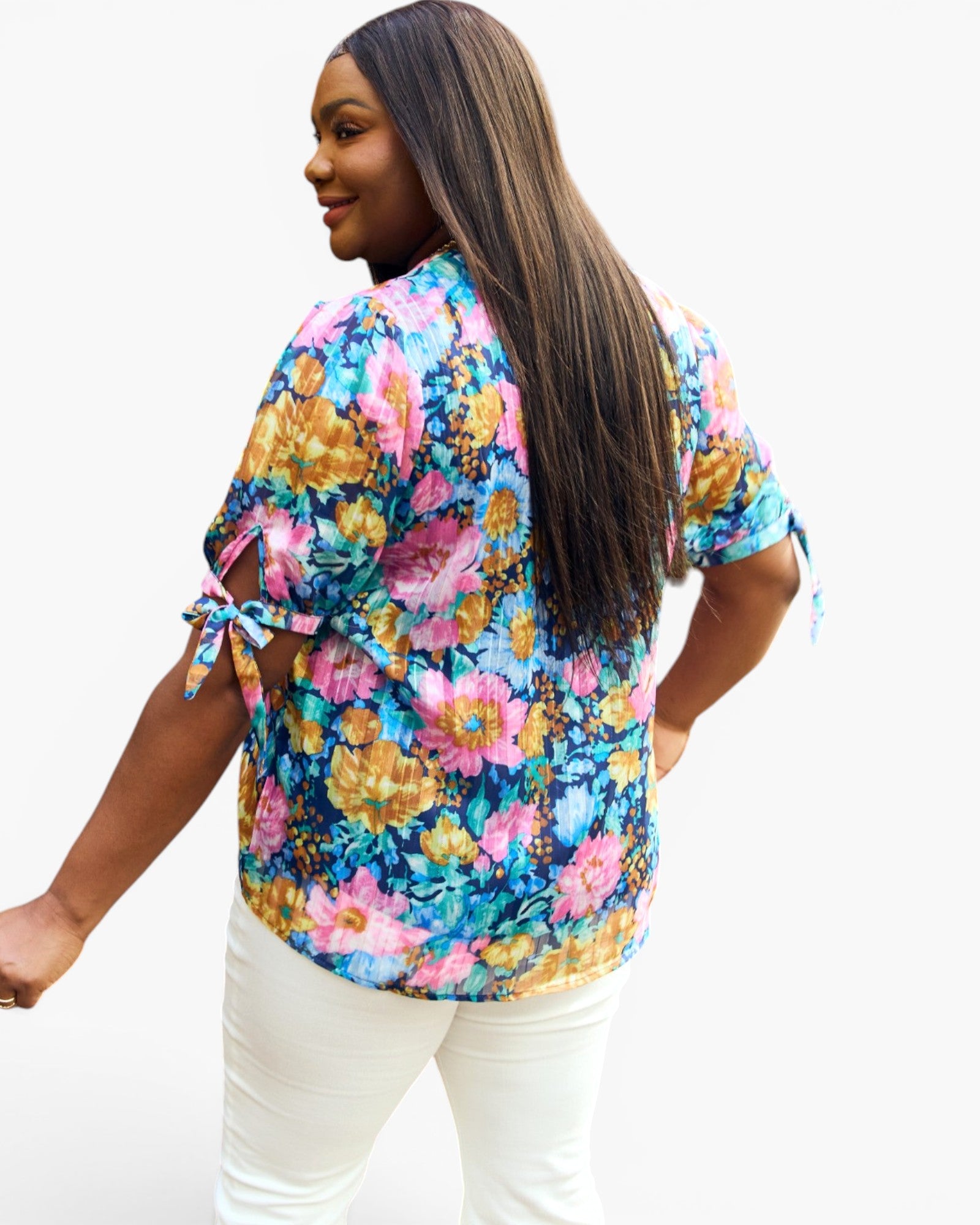 A woman wearing a colorful floral short-sleeve blouse with tie details on the sleeves, paired with white pants, shown from a back-three-quarter angle against a plain white background.
