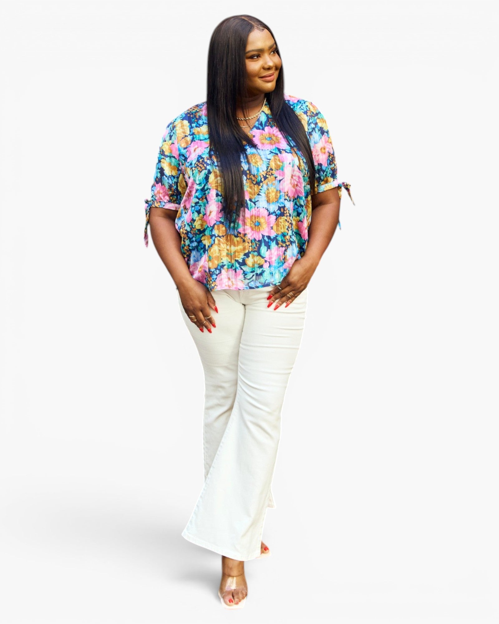 A woman modeling a short-sleeve floral blouse with tie details at the cuffs and white flared pants, standing against a plain light gray studio background.