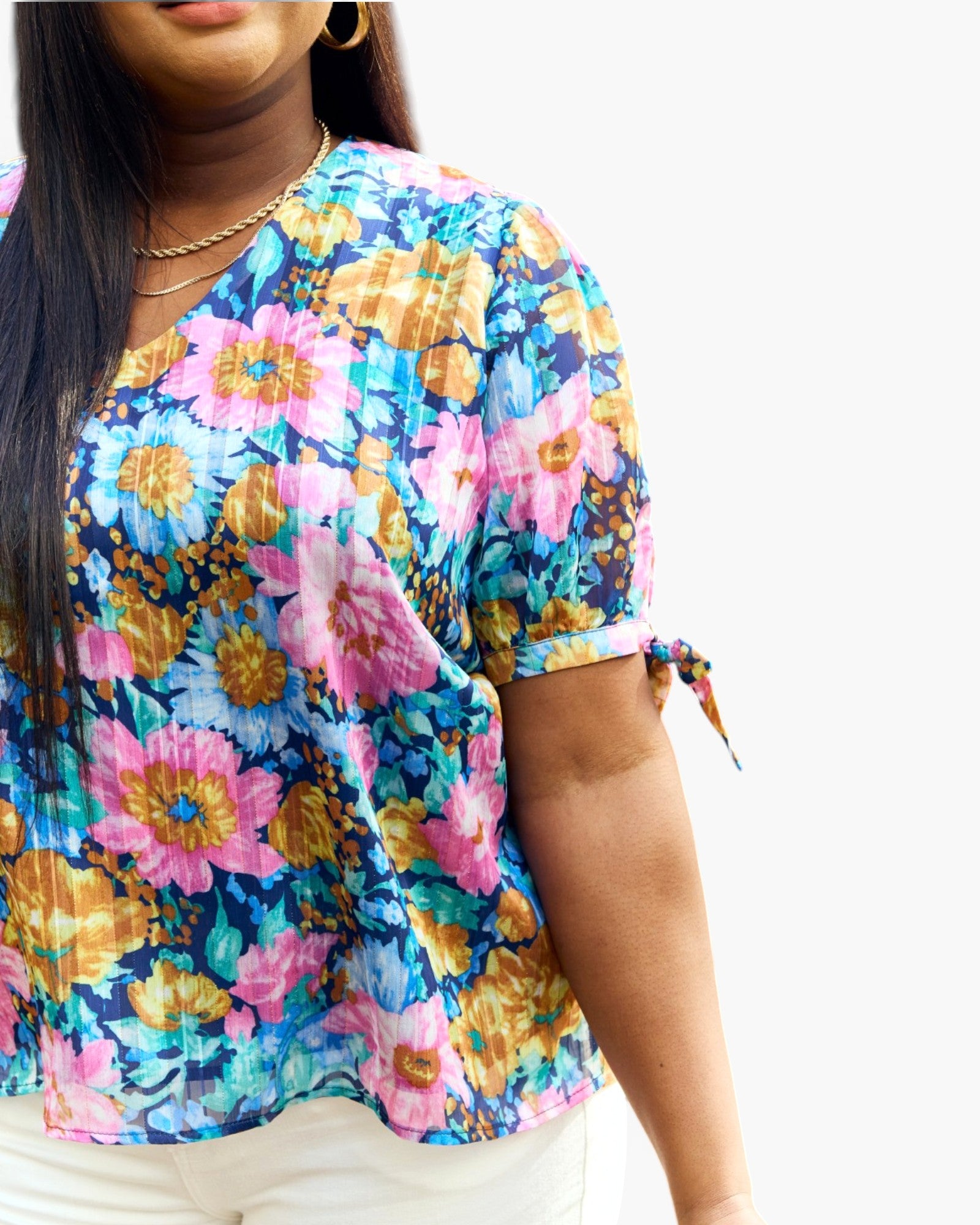 A woman wearing a short-sleeve V-neck blouse with a colorful floral print in pink, blue, yellow, and orange, paired with white pants against a plain white background.