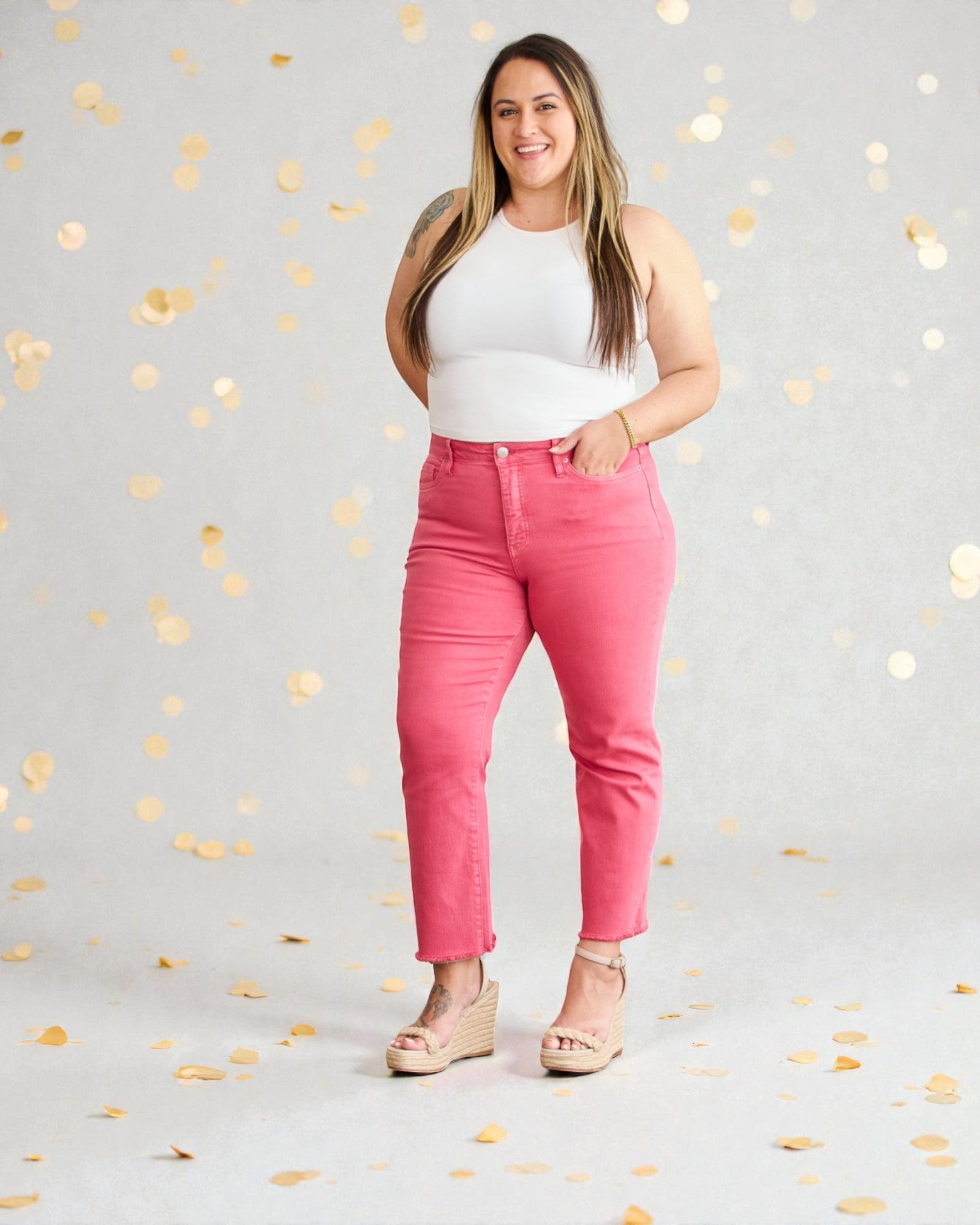 Woman wearing pink jeans and a white top against a gray background with gold confetti.