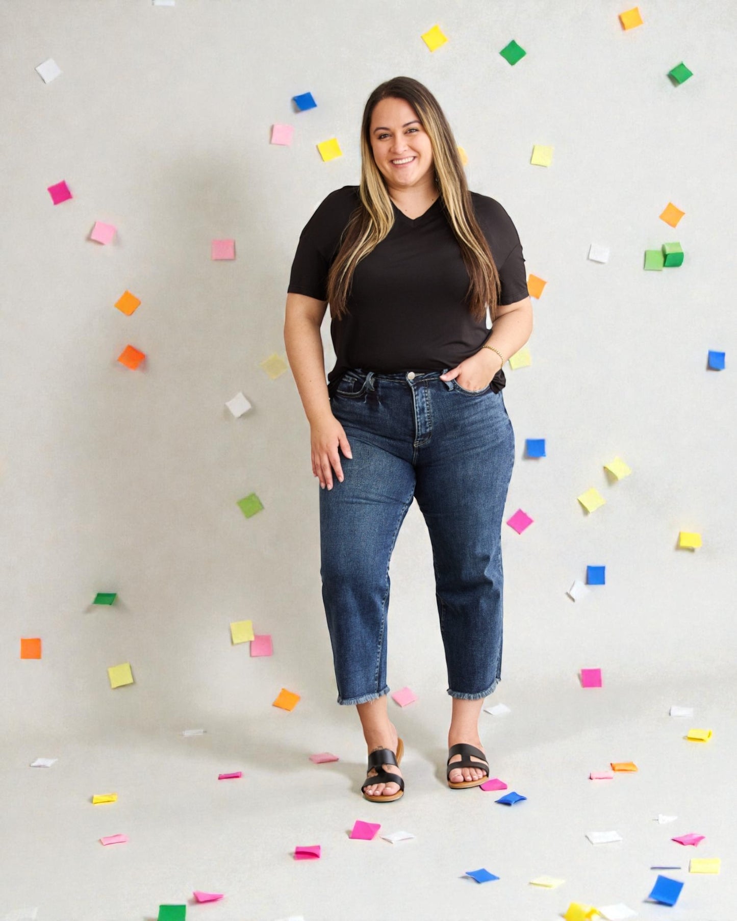 Woman wearing a black t-shirt and blue jeans standing against a light gray background with colorful confetti.