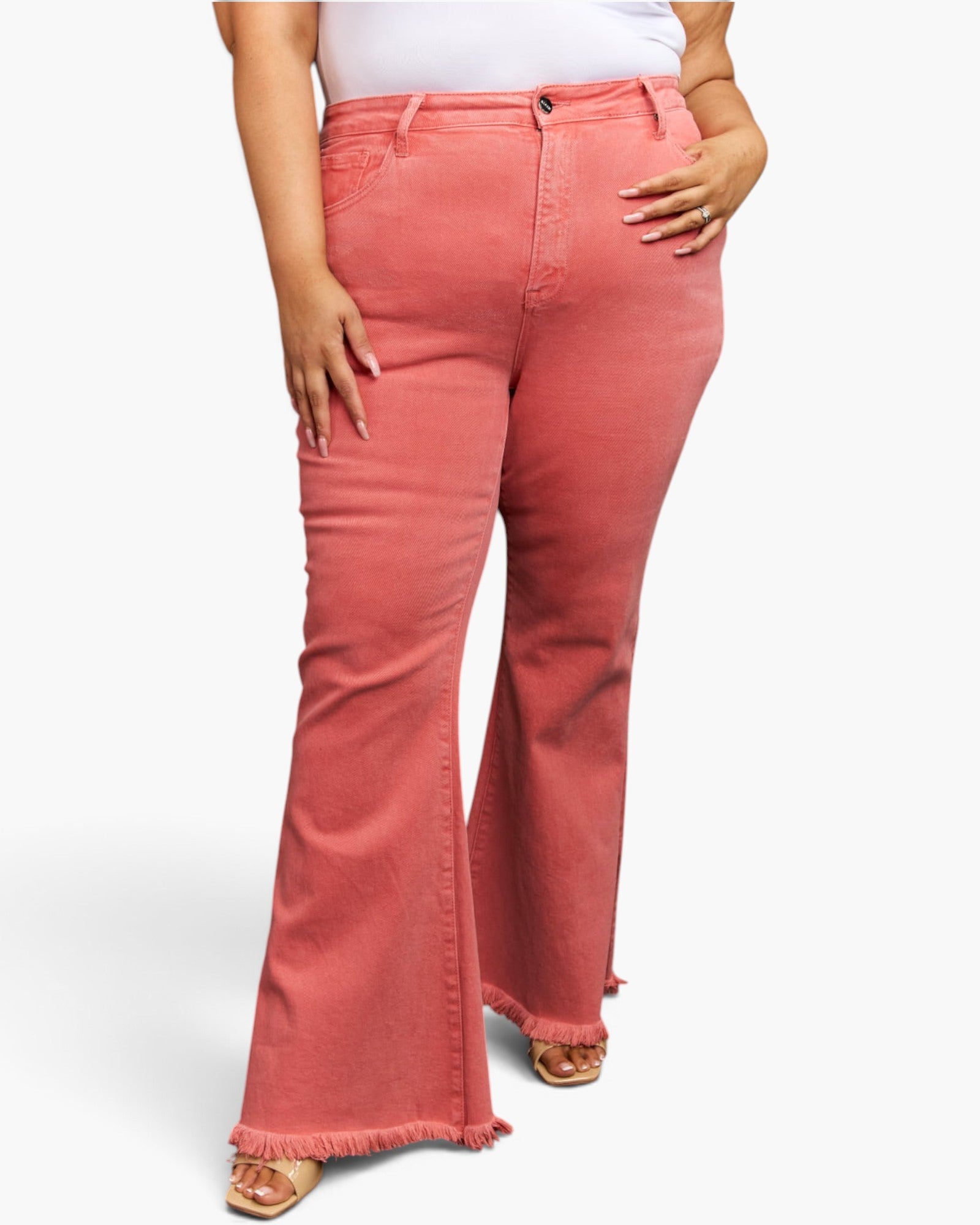 A model wearing high-waisted, flared denim jeans in a washed coral pink color, paired with a white top and tan sandals, shown from the waist down on a light gray background.