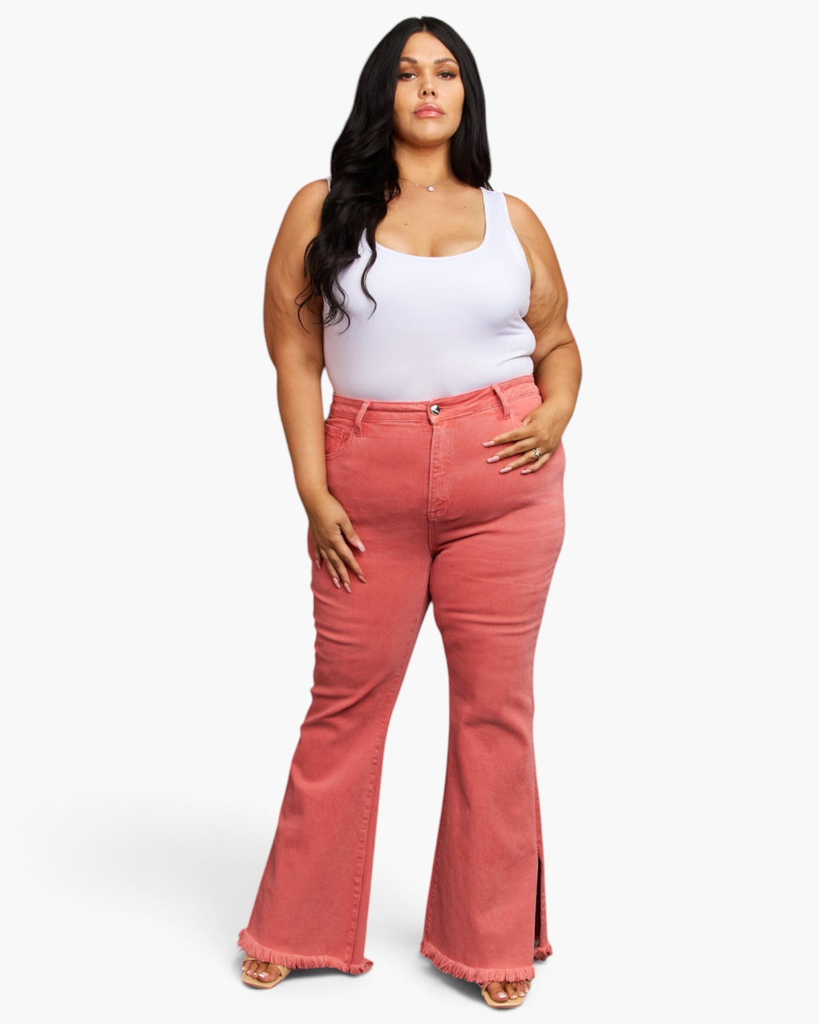 A plus-size model wearing a white tank top and high-waisted, flared coral pink denim jeans with side slits, standing against a light gray studio background.