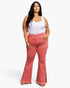 A plus-size model wearing a white tank top and high-waisted, flared coral pink denim jeans with side slits, standing against a light gray studio background.