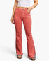 A model wearing a pair of high-waisted, flared denim jeans in a washed coral pink color, paired with a white crop top and brown platform sandals, against a light gray studio background.