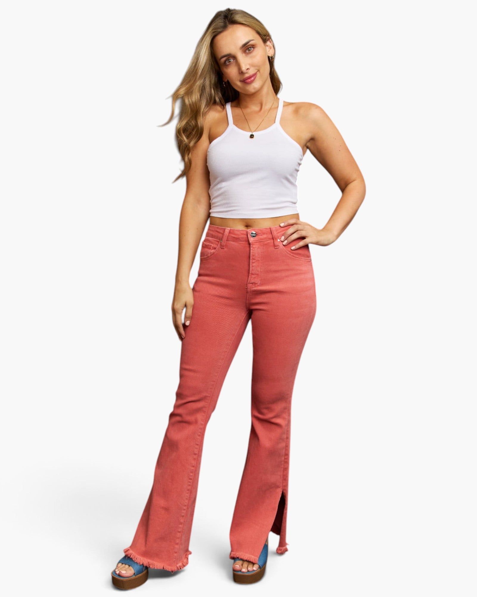 A woman modeling a pair of high-waisted, flared denim jeans in a dusty rose/coral color, paired with a white ribbed tank top and blue platform sandals, against a plain white studio background.