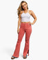 A woman modeling a pair of high-waisted, flared denim jeans in a dusty rose/coral color, paired with a white ribbed tank top and blue platform sandals, against a plain white studio background.