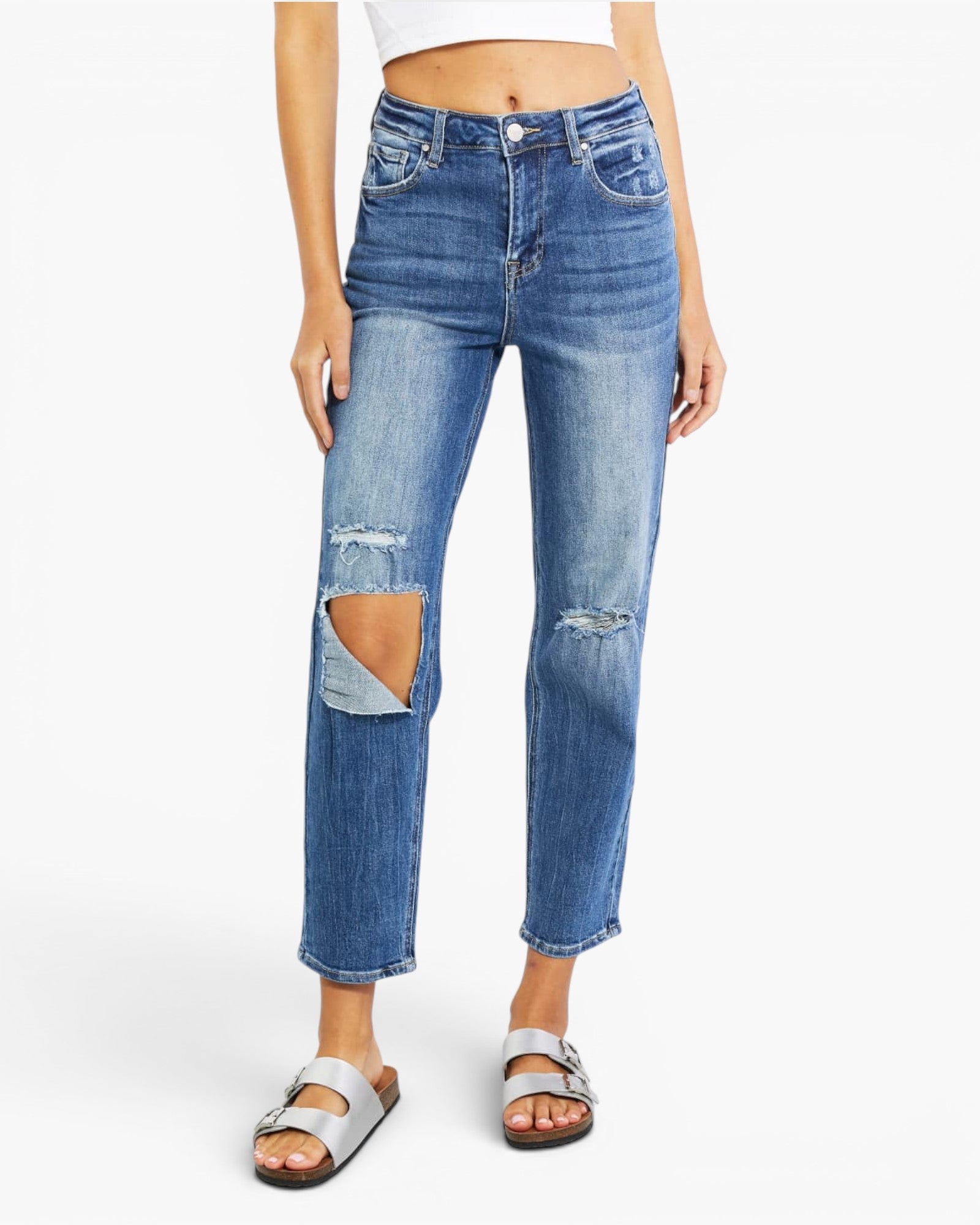 A model wearing medium-wash blue cropped jeans with distressed rips at the knees, paired with a white crop top and silver slide sandals, against a plain white background.