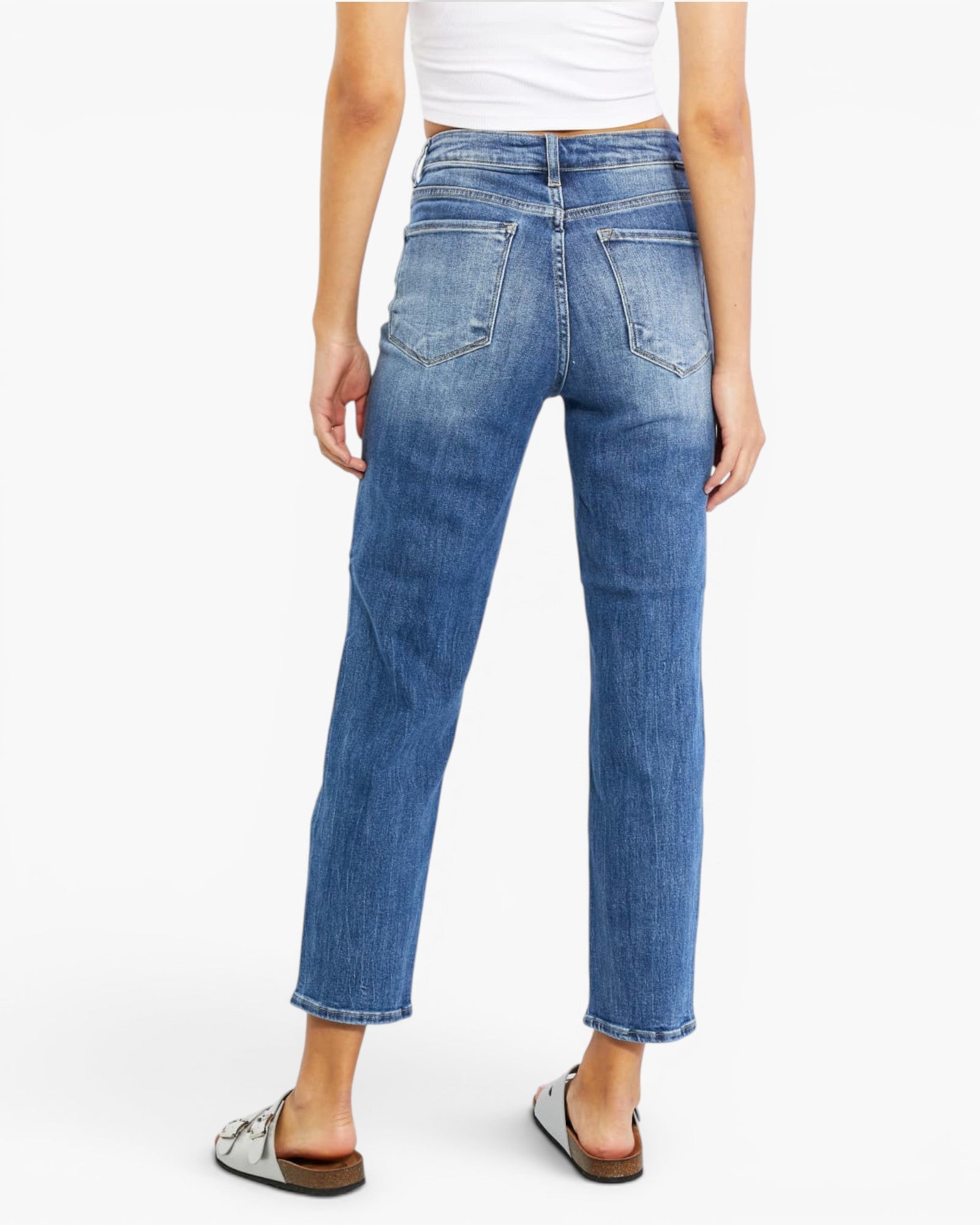 A person shown from the back wearing medium-wash blue cropped jeans with a white cropped top and brown slide sandals against a light gray studio background.