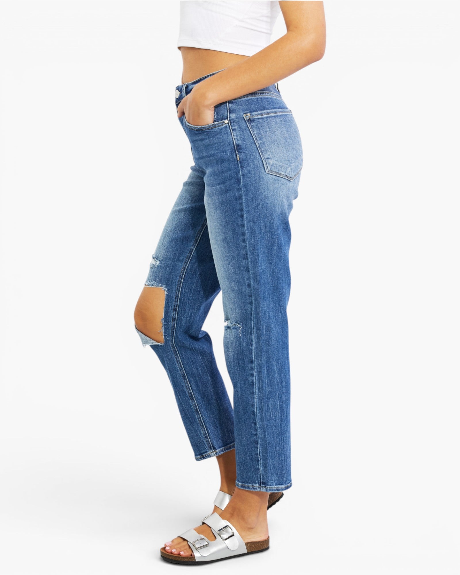A side profile view of a person wearing medium-wash blue distressed jeans with ripped details on the knees, styled with a white crop top and white slide sandals against a plain white background.
