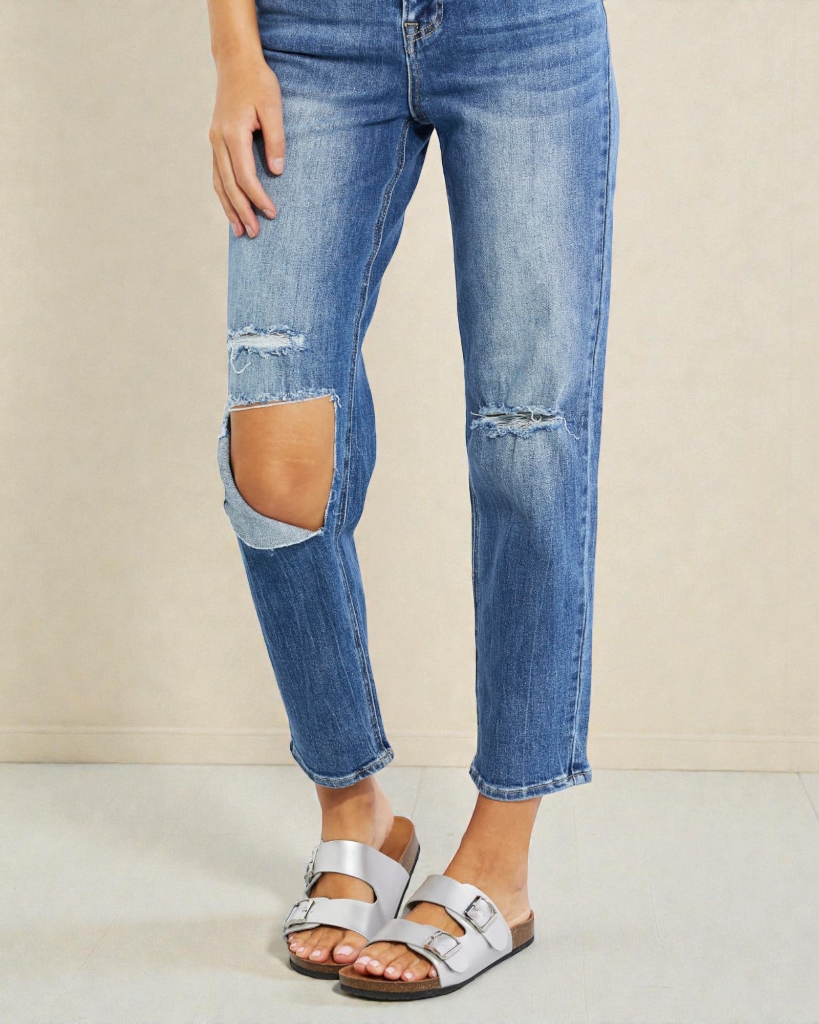 Person wearing ripped blue jeans and silver sandals on a beige background