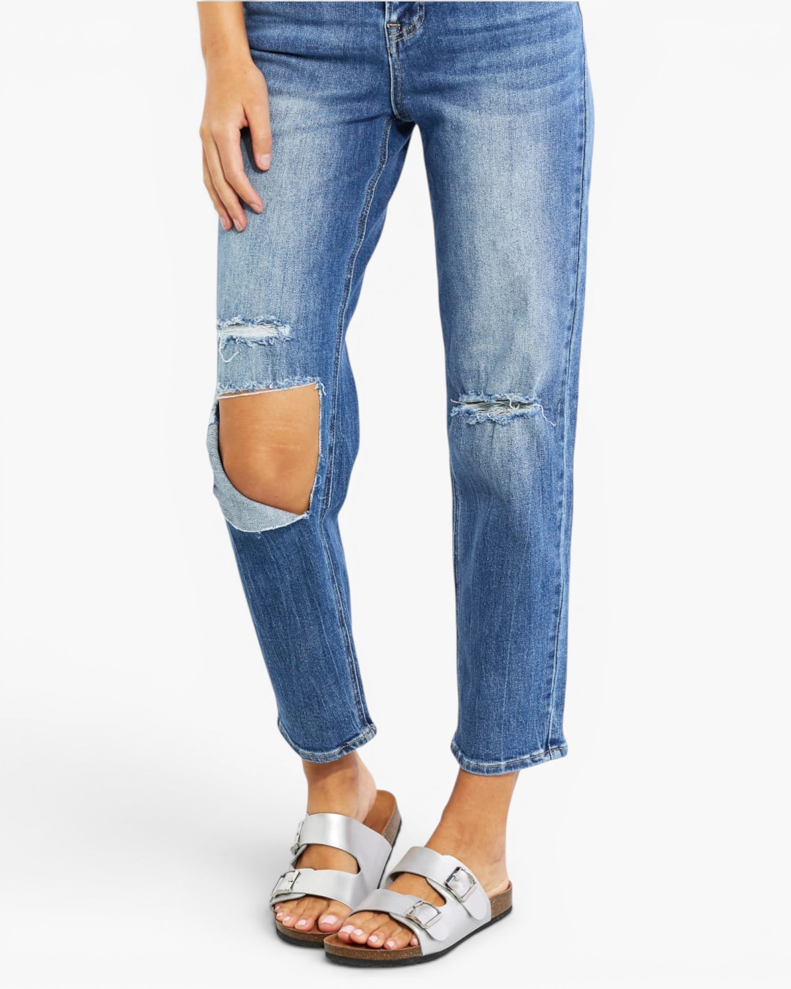 A person wearing medium-wash blue distressed jeans with large ripped knee patches and silver double-strap sandals, shown from the waist down on a light gray studio background.