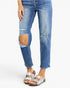 A person wearing medium-wash blue distressed jeans with large ripped knee patches and silver double-strap sandals, shown from the waist down on a light gray studio background.