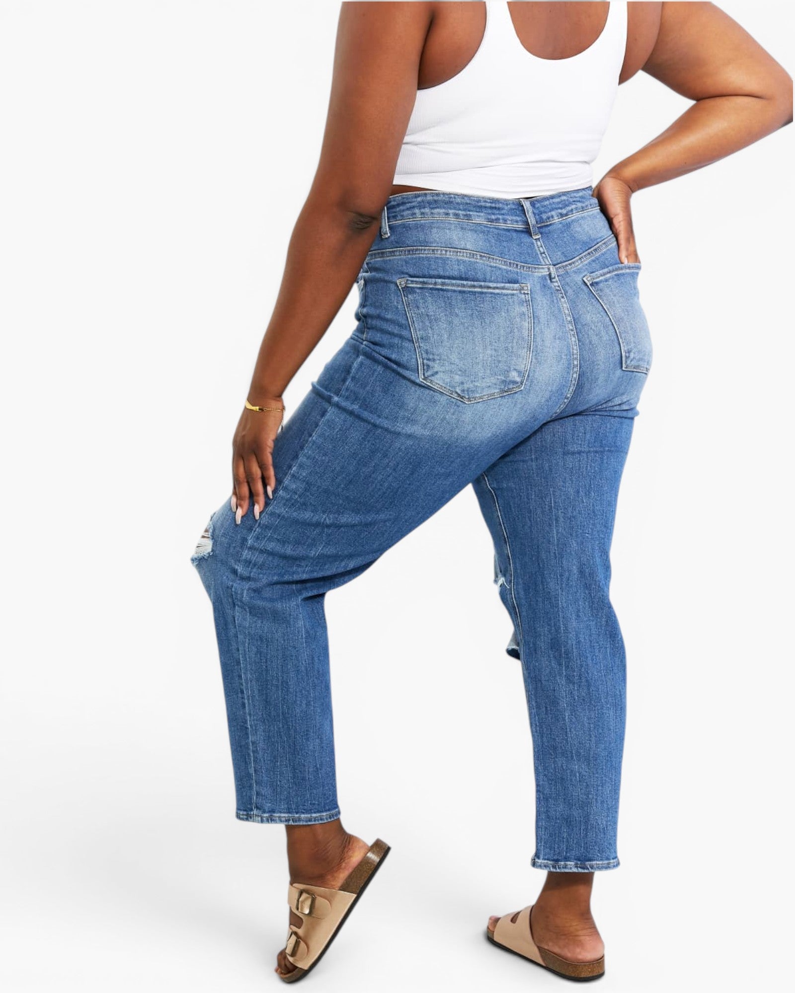 A model from the back wearing high-waisted, cropped blue denim jeans with a white tank top and beige slide sandals, set against a plain white studio background.