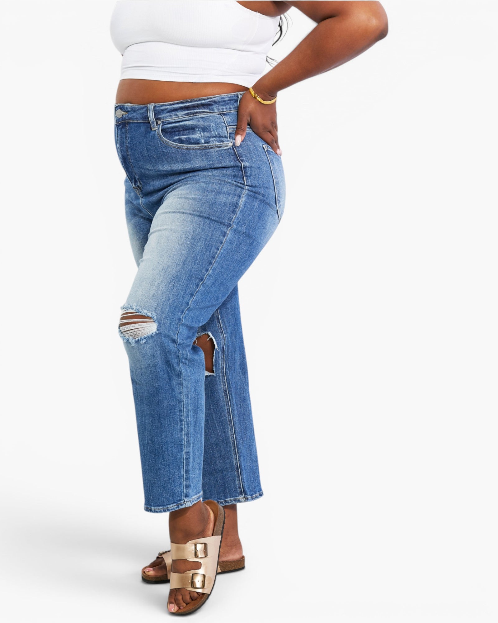 A model wearing high-waisted, cropped blue denim jeans with distressed rips and a side slit, styled with a white crop top and beige double-strap sandals against a light gray studio background.