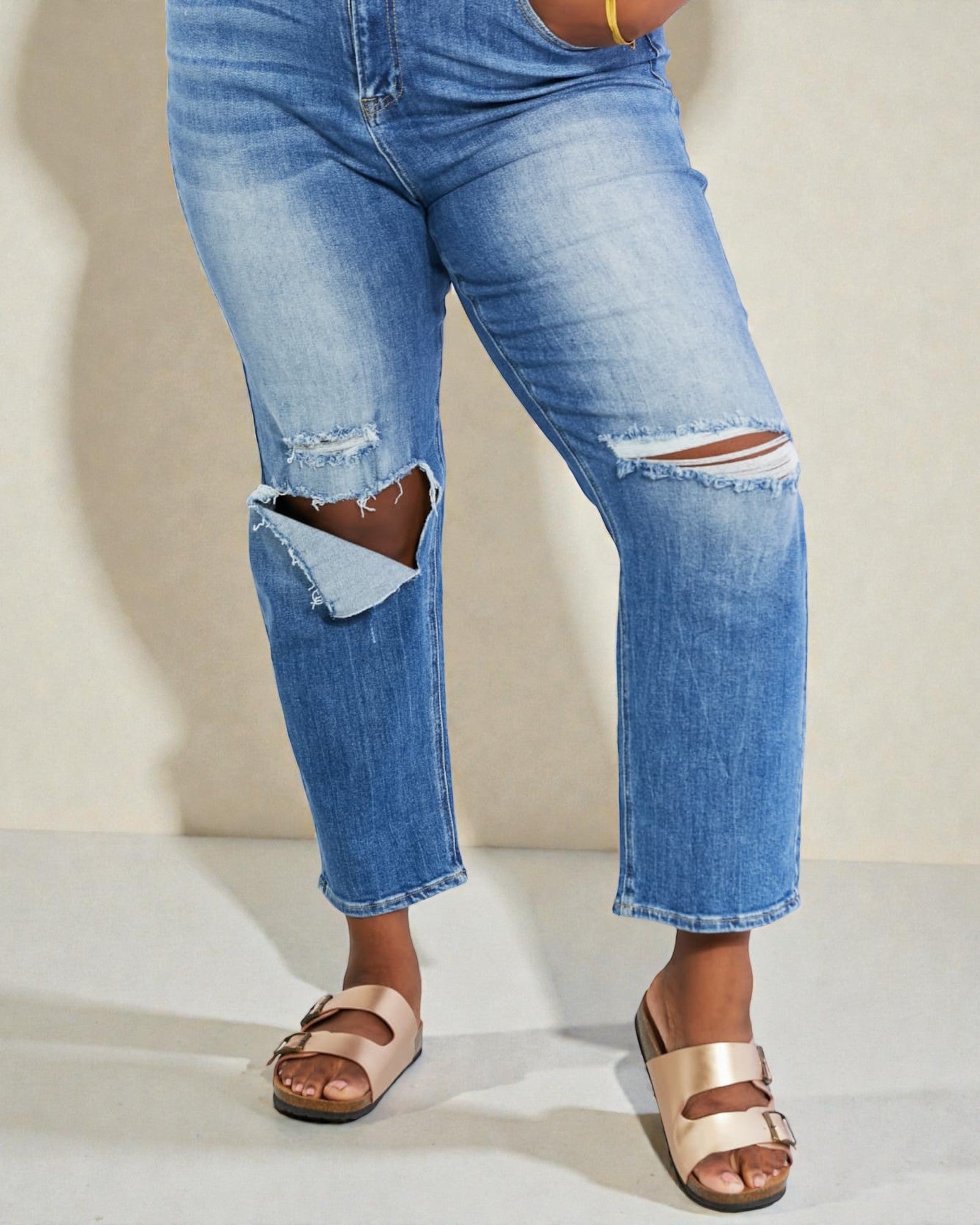 Person wearing ripped blue jeans and beige sandals on a neutral background