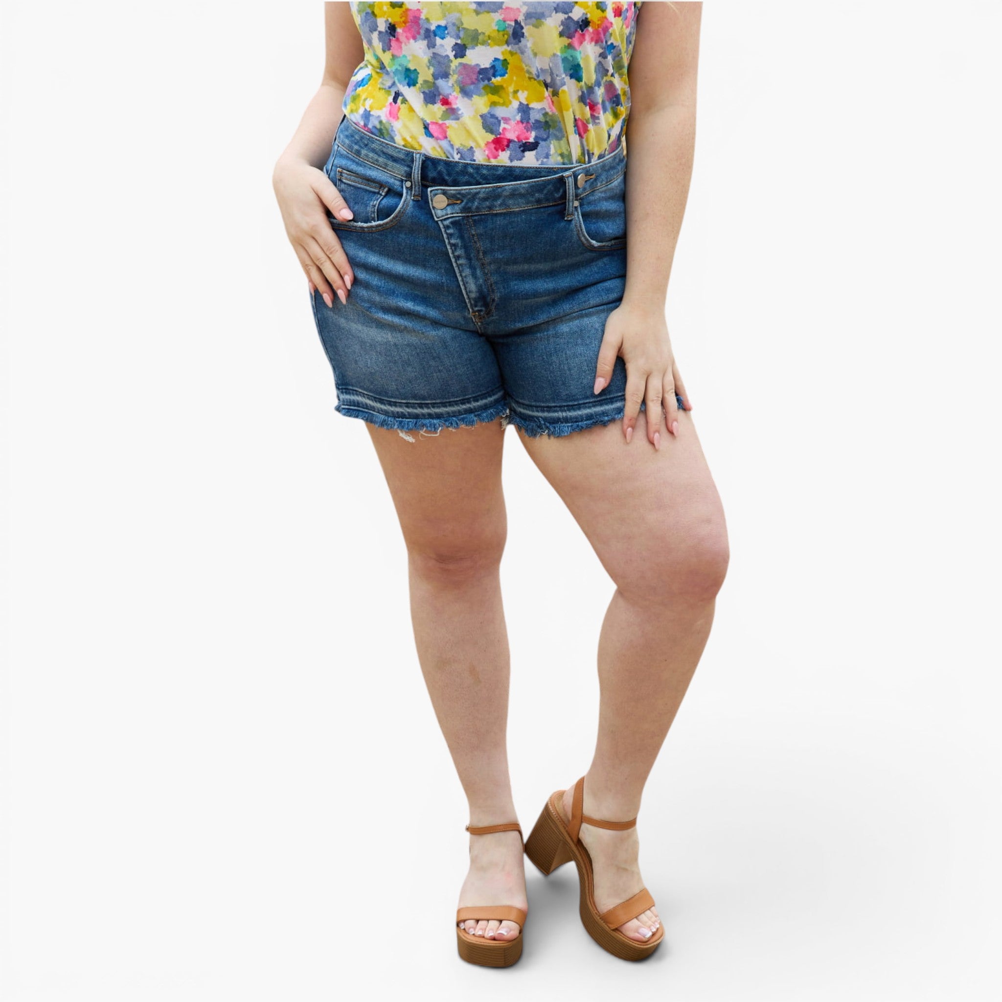 A model wearing medium-wash blue denim cut-off shorts with a frayed hem, paired with a colorful floral top and brown platform sandals, shown from the waist down on a light gray background.