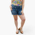A model wearing medium-wash blue denim cut-off shorts with a frayed hem, paired with a colorful floral top and brown platform sandals, shown from the waist down on a light gray background.