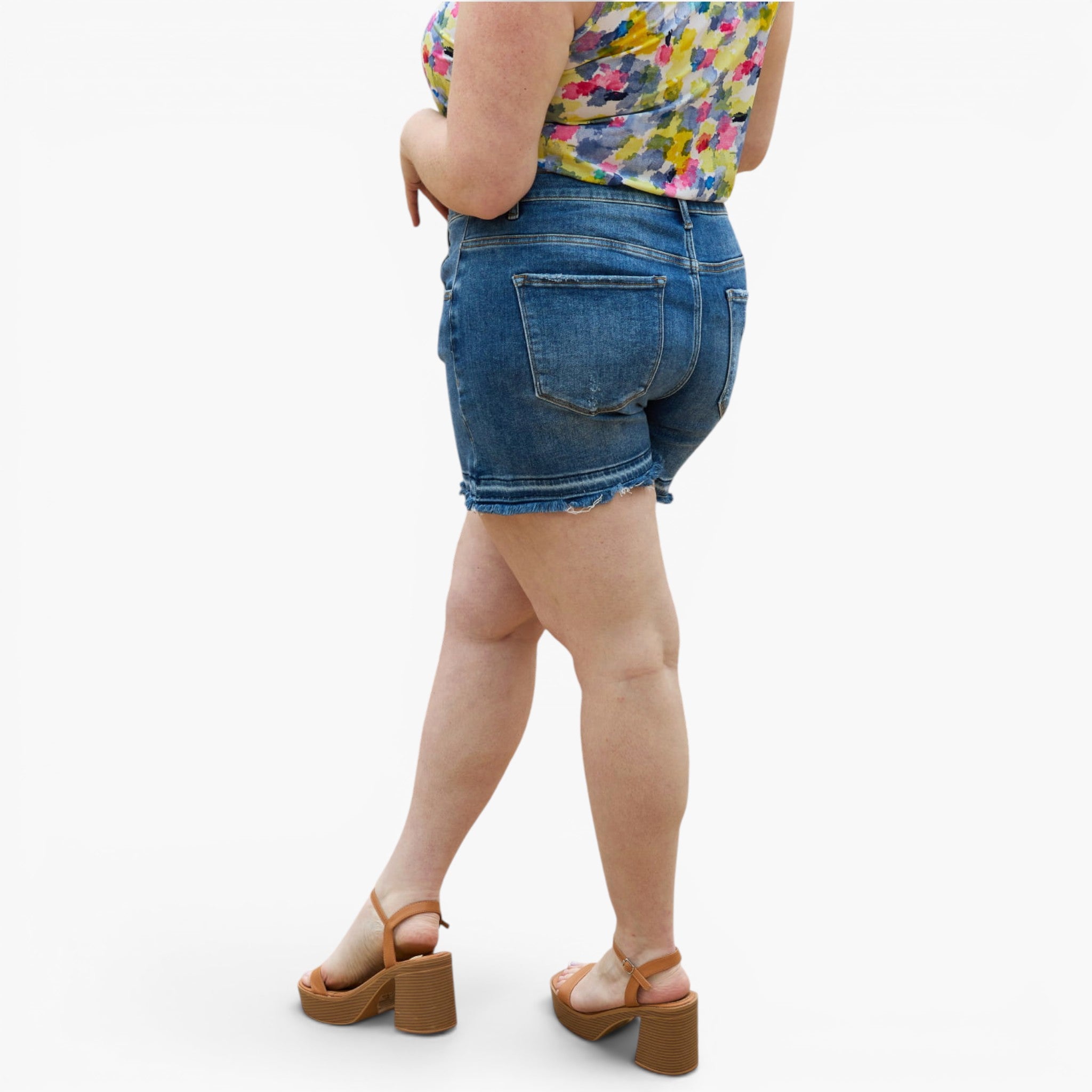 A person shown from the back wearing blue denim shorts, a colorful floral top, and tan platform sandals against a plain light gray studio background.