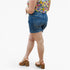 A person shown from the back wearing blue denim shorts, a colorful floral top, and tan platform sandals against a plain light gray studio background.