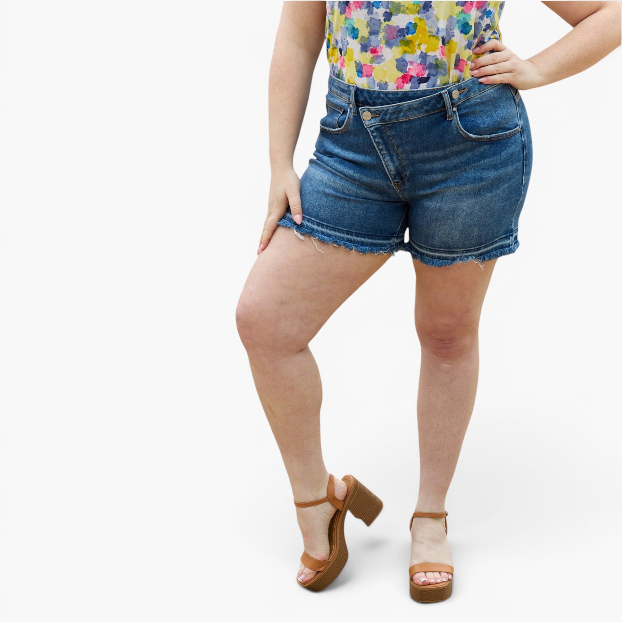 A model wearing blue denim shorts with a raw, frayed hem, paired with a colorful floral top and brown platform sandals, shown against a plain white studio background.