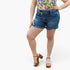 A model wearing blue denim shorts with a raw, frayed hem, paired with a colorful floral top and brown platform sandals, shown against a plain white studio background.