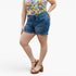 A model wearing medium-wash blue denim shorts with a frayed hem, paired with a colorful floral top and brown platform sandals, shown against a plain white studio background.