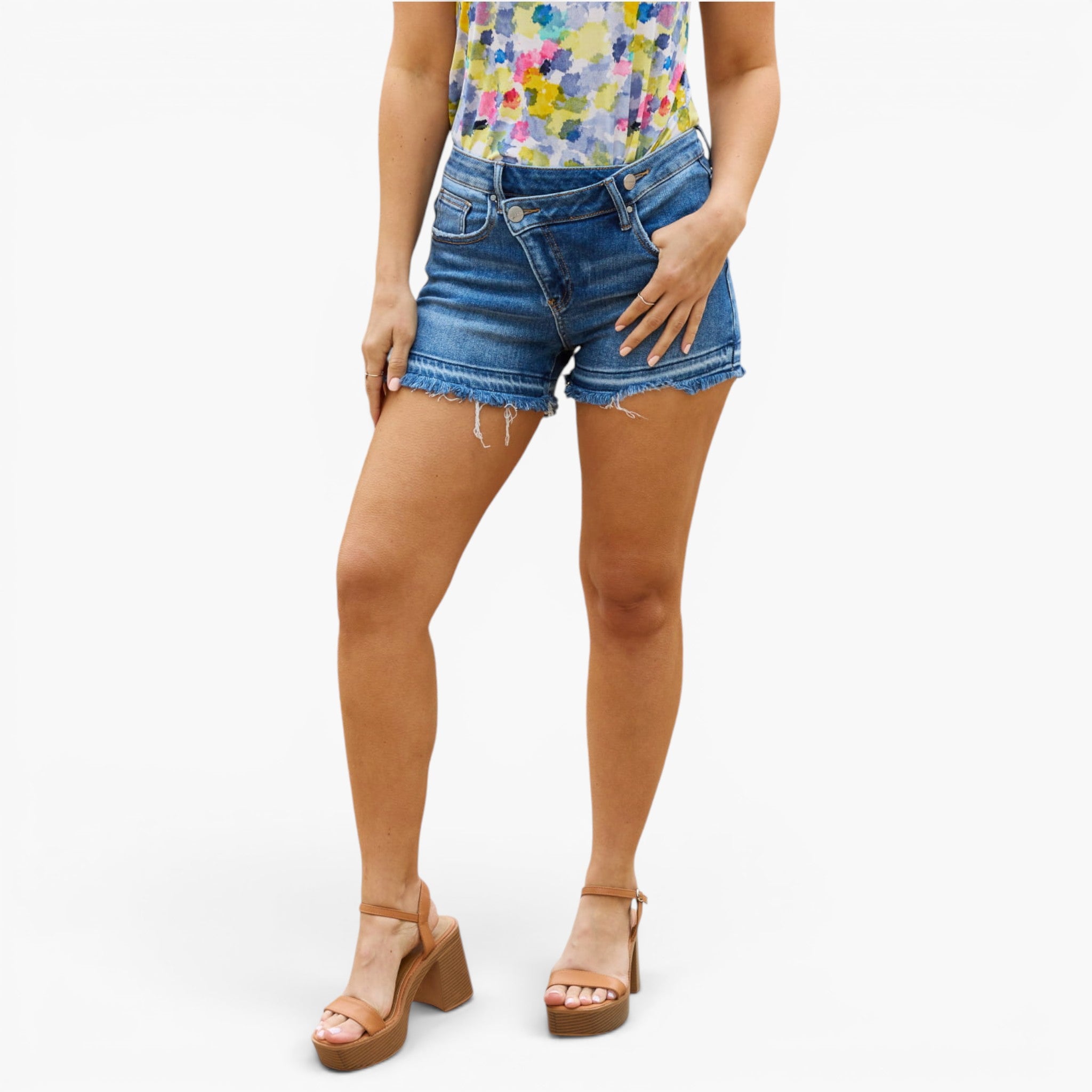 A model wearing medium-wash blue denim cut-off shorts with a frayed hem, paired with a colorful floral top and brown platform sandals, against a plain white background.