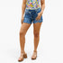A model wearing medium-wash blue denim cut-off shorts with a frayed hem, paired with a colorful floral top and brown platform sandals, against a plain white background.