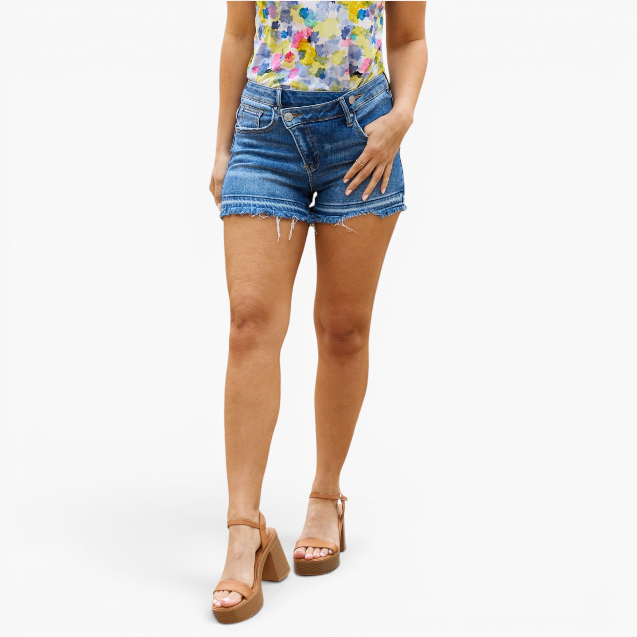 A model wearing medium-wash blue denim cut-off shorts with a raw frayed hem, paired with a colorful floral top and brown platform sandals, shown against a plain white studio background.