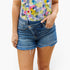 A person wearing medium-wash blue denim cutoff shorts with a frayed hem and a colorful floral print top, shown from the chest to the upper thighs against a light gray studio background.
