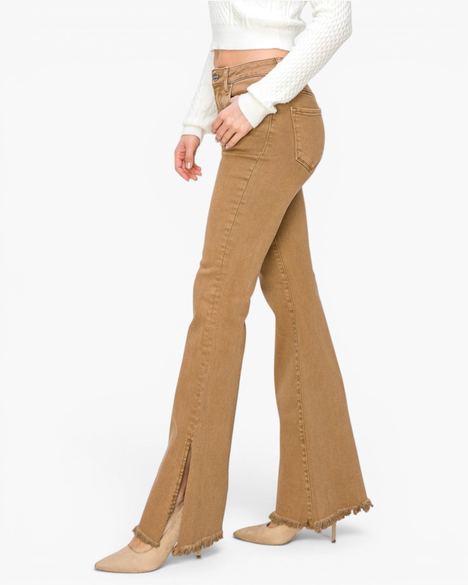 A model is shown from the waist down wearing tan flare jeans with fringe detailing at the hem and side slits, paired with a white textured long-sleeve crop top and beige pointed-toe heels against a plain white background.