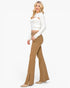 A woman modeling a white ribbed long-sleeve crop top with cutout details and brown flared jeans, paired with beige pointed-toe heels, against a plain white background.