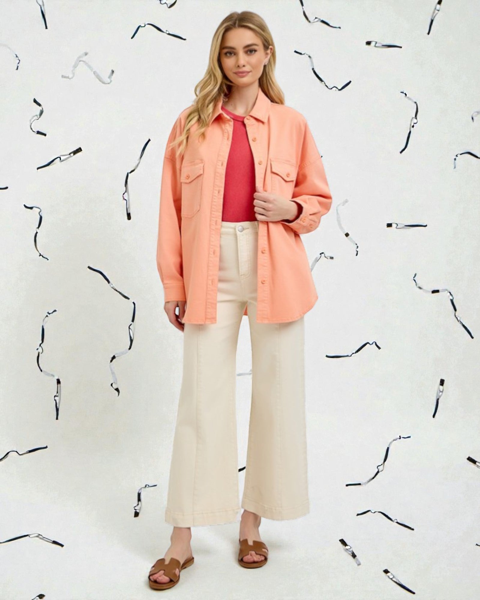Woman wearing a coral shirt over a pink top with beige pants against a white background with black abstract patterns.