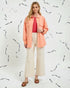 Woman wearing a coral shirt over a pink top with beige pants against a white background with black abstract patterns.