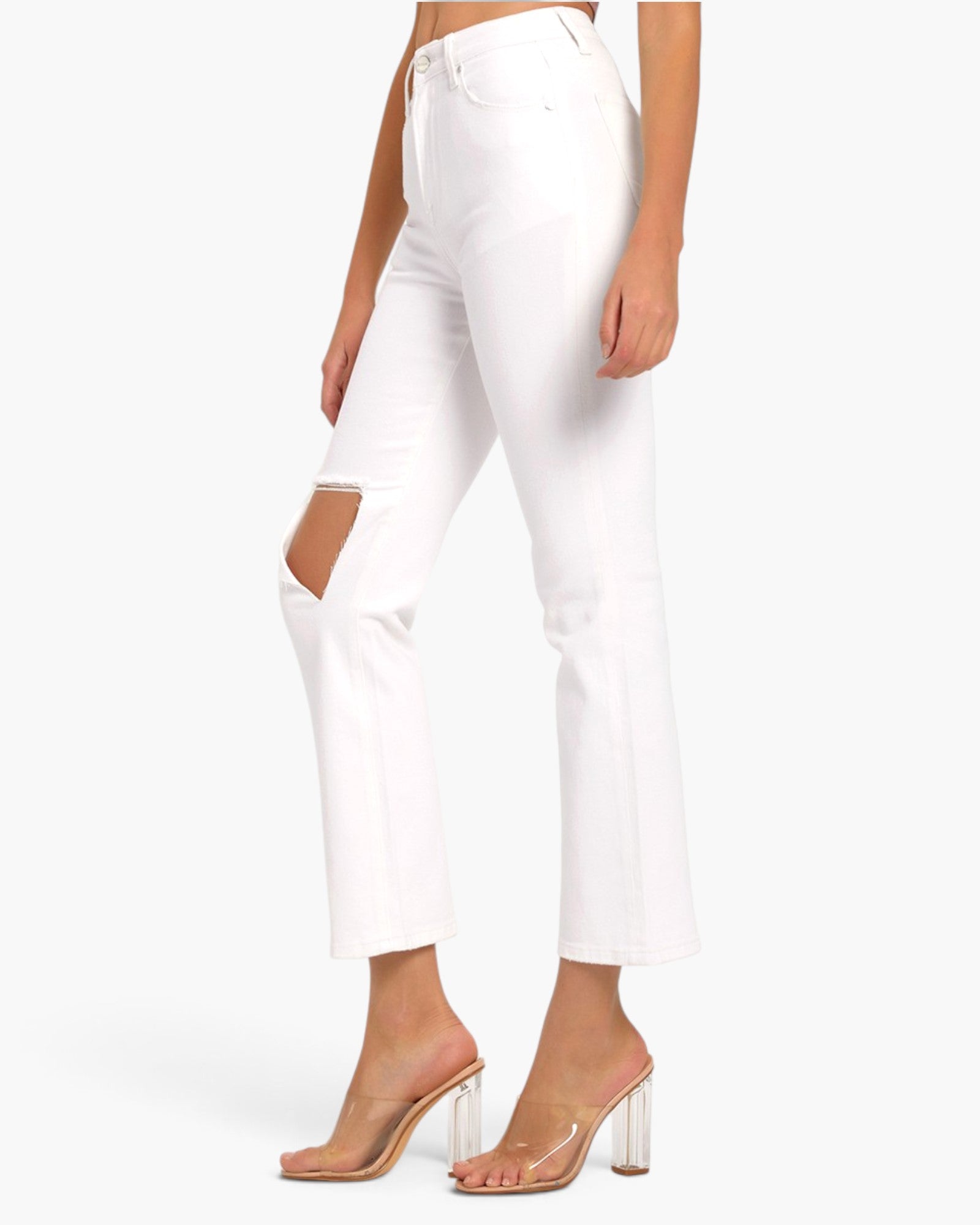 White cropped jeans with a diagonal cutout on the left leg, modeled with clear high-heel mules against a plain white background.