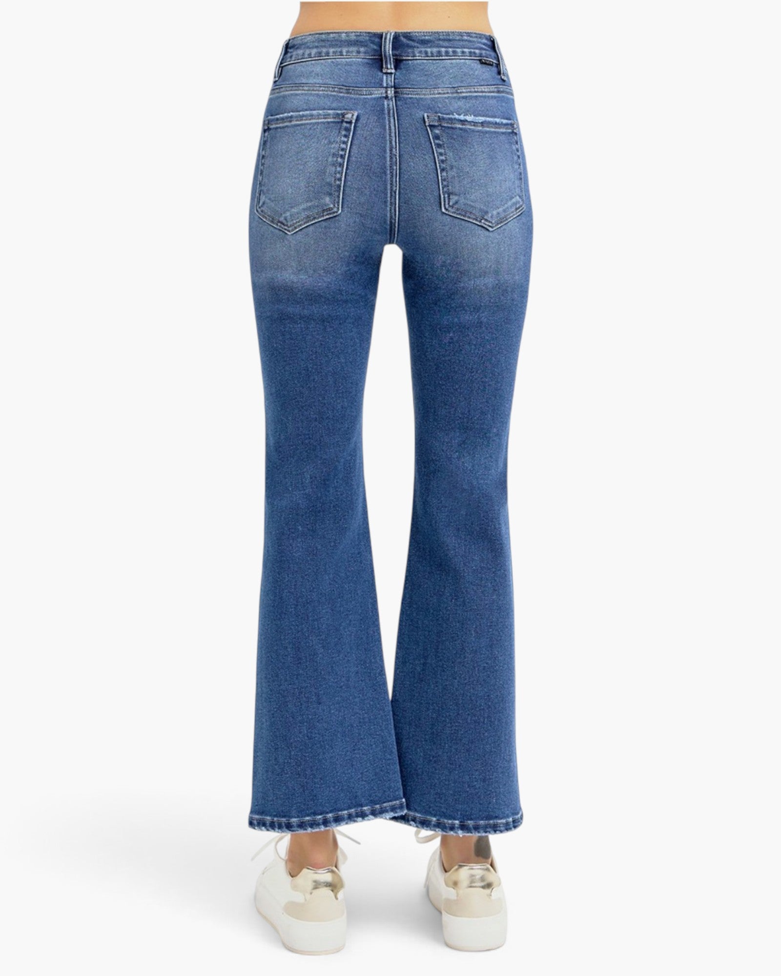 Rear view of a person wearing blue flare jeans and white sneakers against a plain light gray studio background.