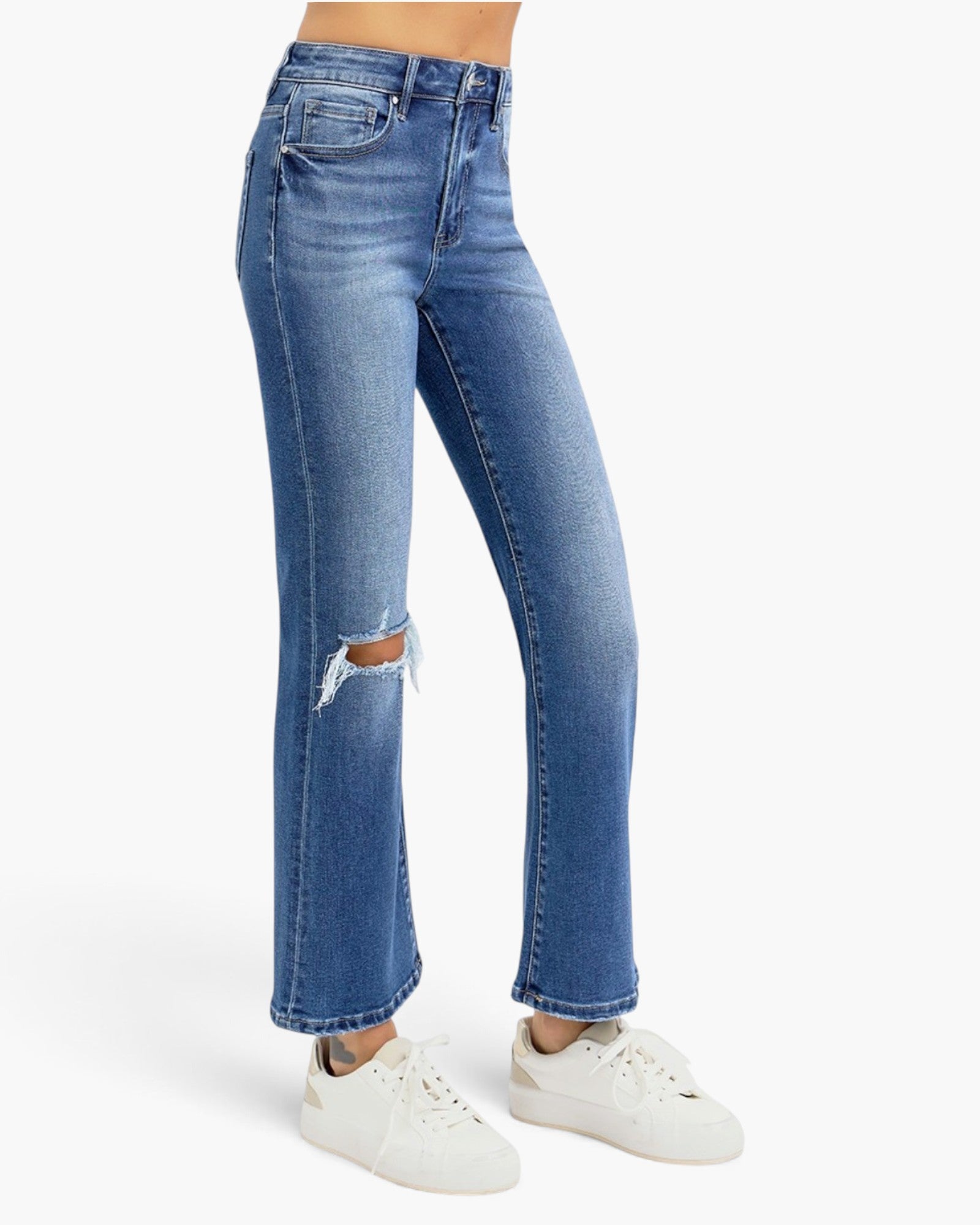 A model wearing a pair of high-waisted, straight-leg blue jeans with a distressed rip on the left knee, paired with white sneakers against a light gray studio background.