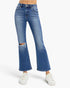 A person wearing medium-wash blue denim flare jeans with a distressed rip at the left knee and white sneakers, shown from the waist down on a light gray studio background.