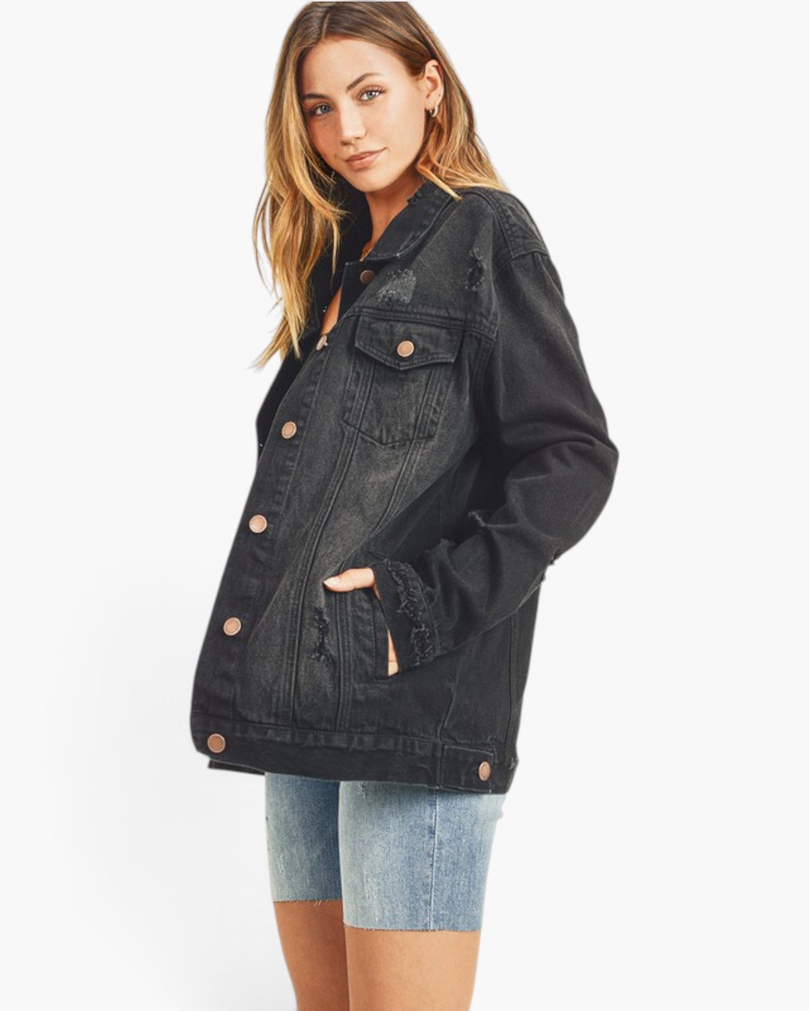 A woman modeling a black washed denim jacket with distressed details and copper-toned buttons, paired with light blue denim shorts, against a plain white studio background.