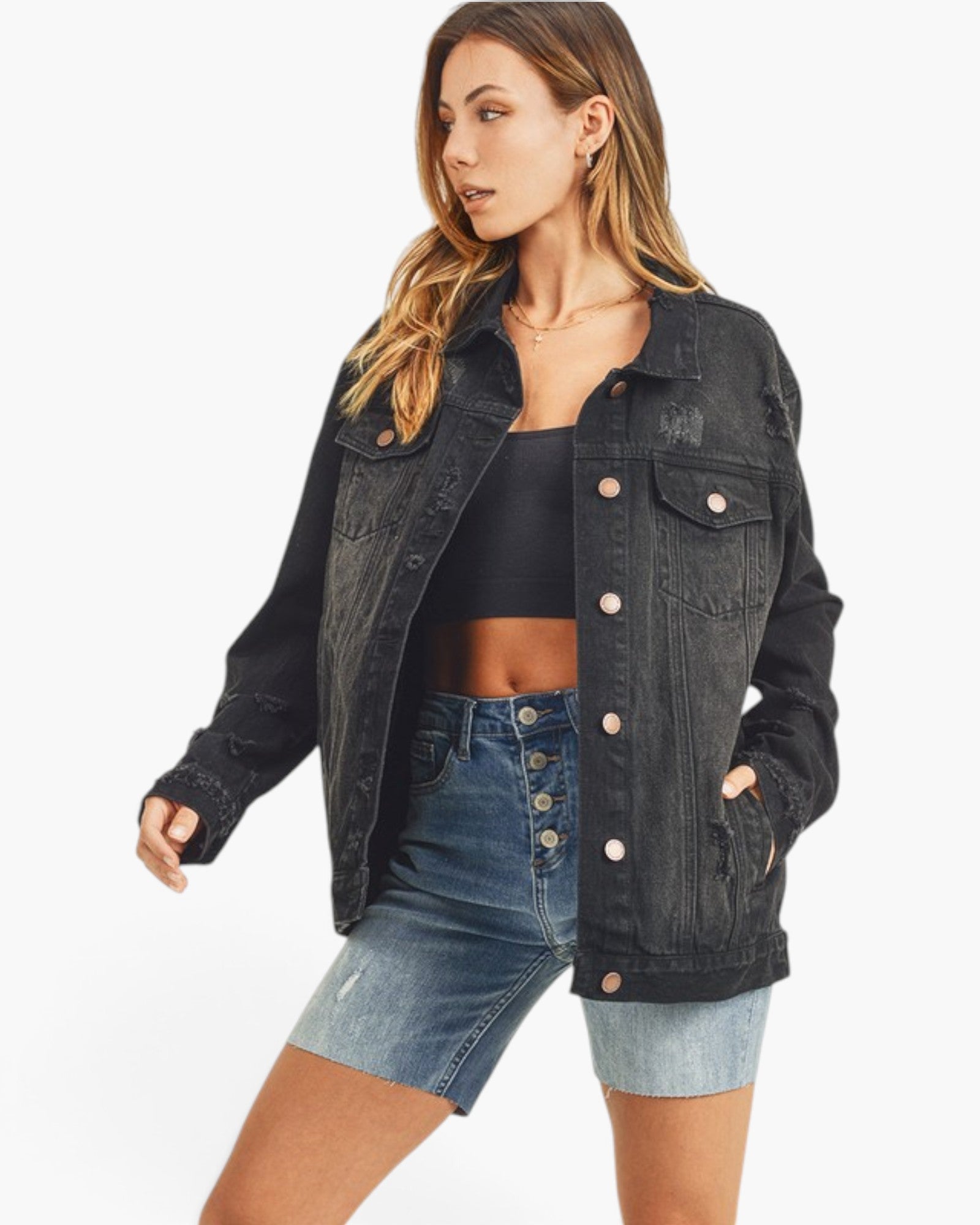 A woman modeling a black distressed denim jacket over a black crop top and light-wash denim shorts against a plain white background.