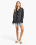 A woman modeling a dark washed denim jacket over a black top, paired with light blue denim shorts and white slide sandals, against a plain white studio background.