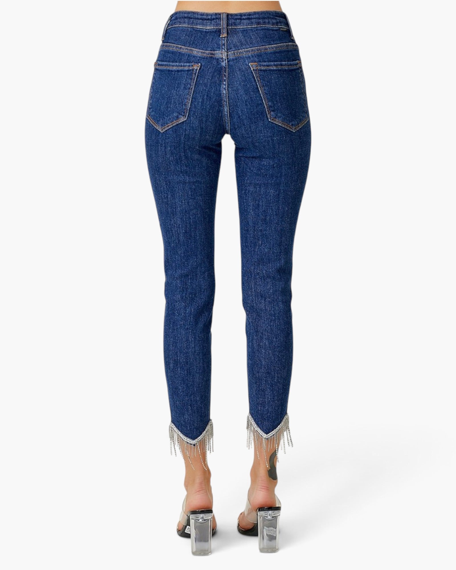 Rear view of a person wearing dark blue denim jeans with frayed hems and clear high-heeled sandals against a light gray studio background.