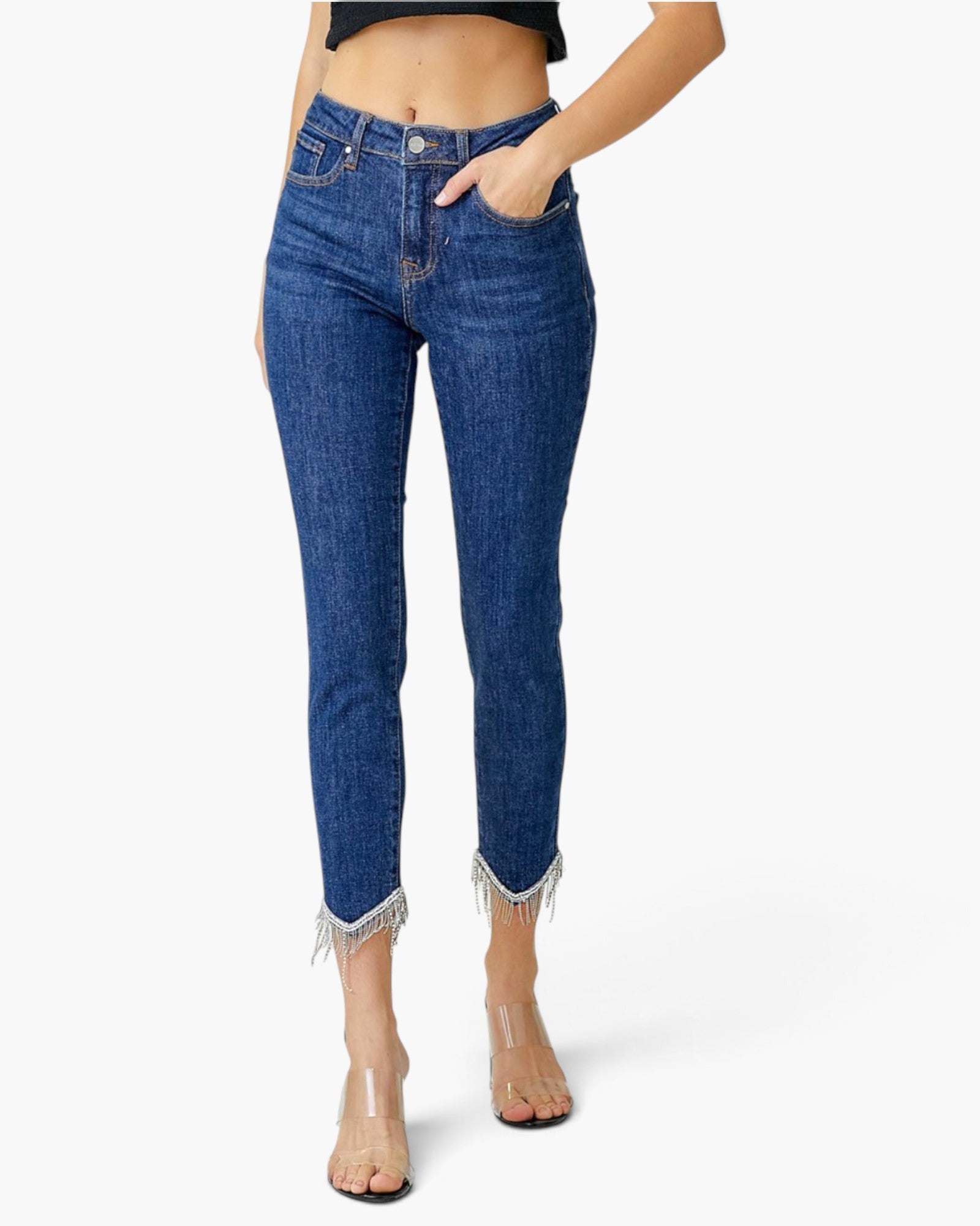 A model wearing a pair of dark blue high-waisted skinny jeans with frayed hems, paired with clear high-heeled sandals against a plain white background.
