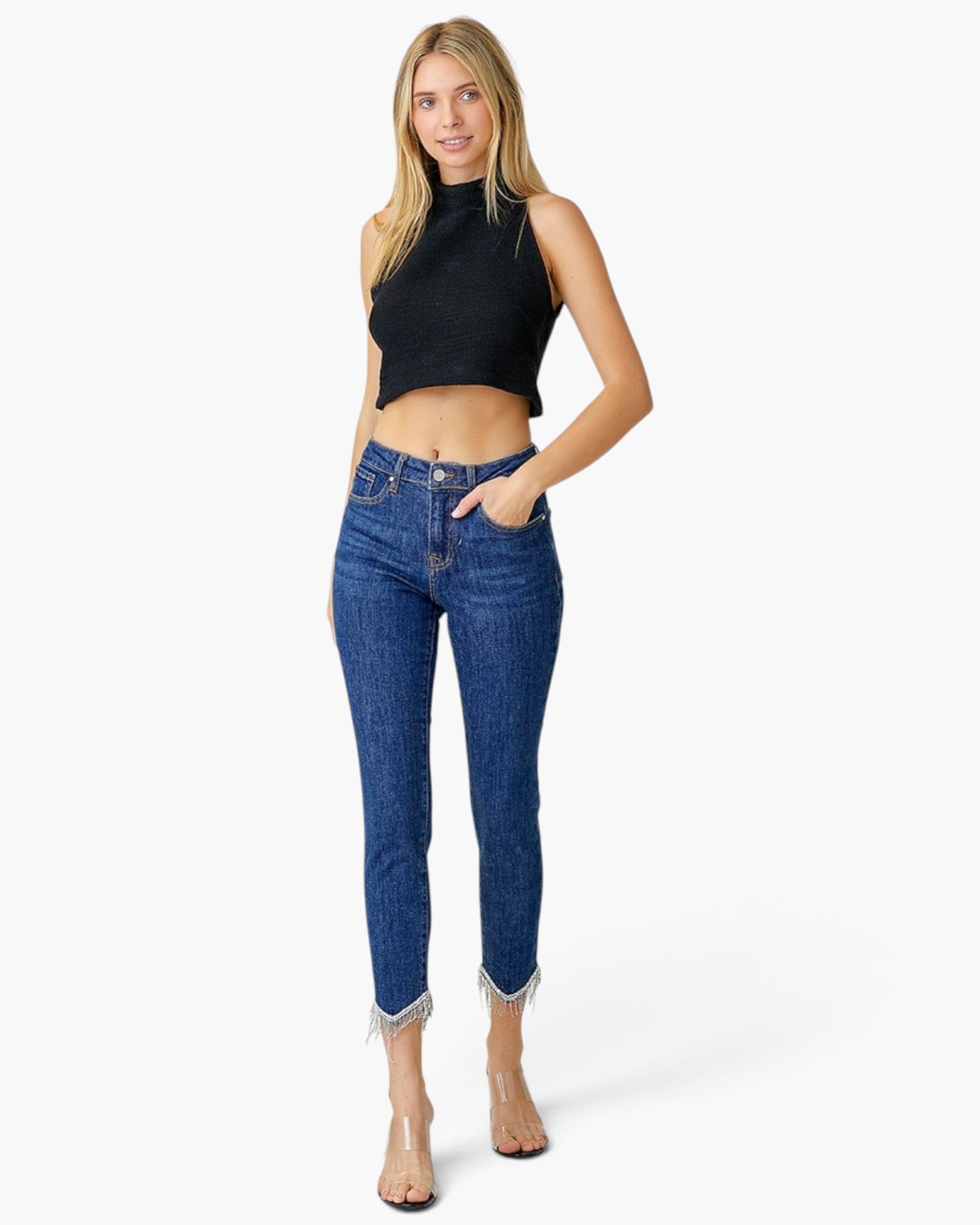 A woman modeling a black sleeveless crop top and blue high-waisted ankle-length jeans with frayed hems, paired with nude high heels, against a plain white studio background.
