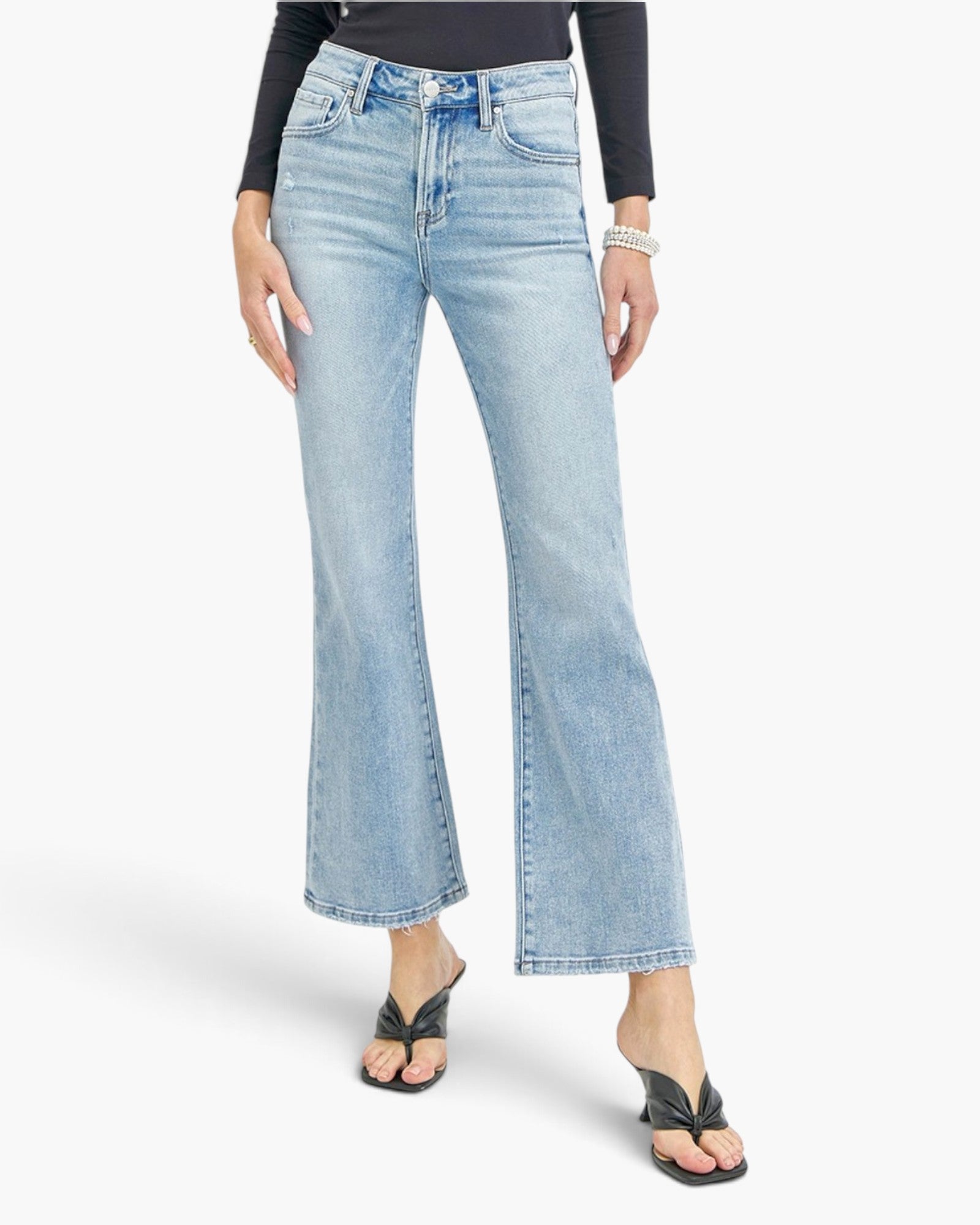 A model wearing a pair of light-wash blue flare jeans, styled with a black long-sleeve top and black strap sandals, against a plain light gray background.