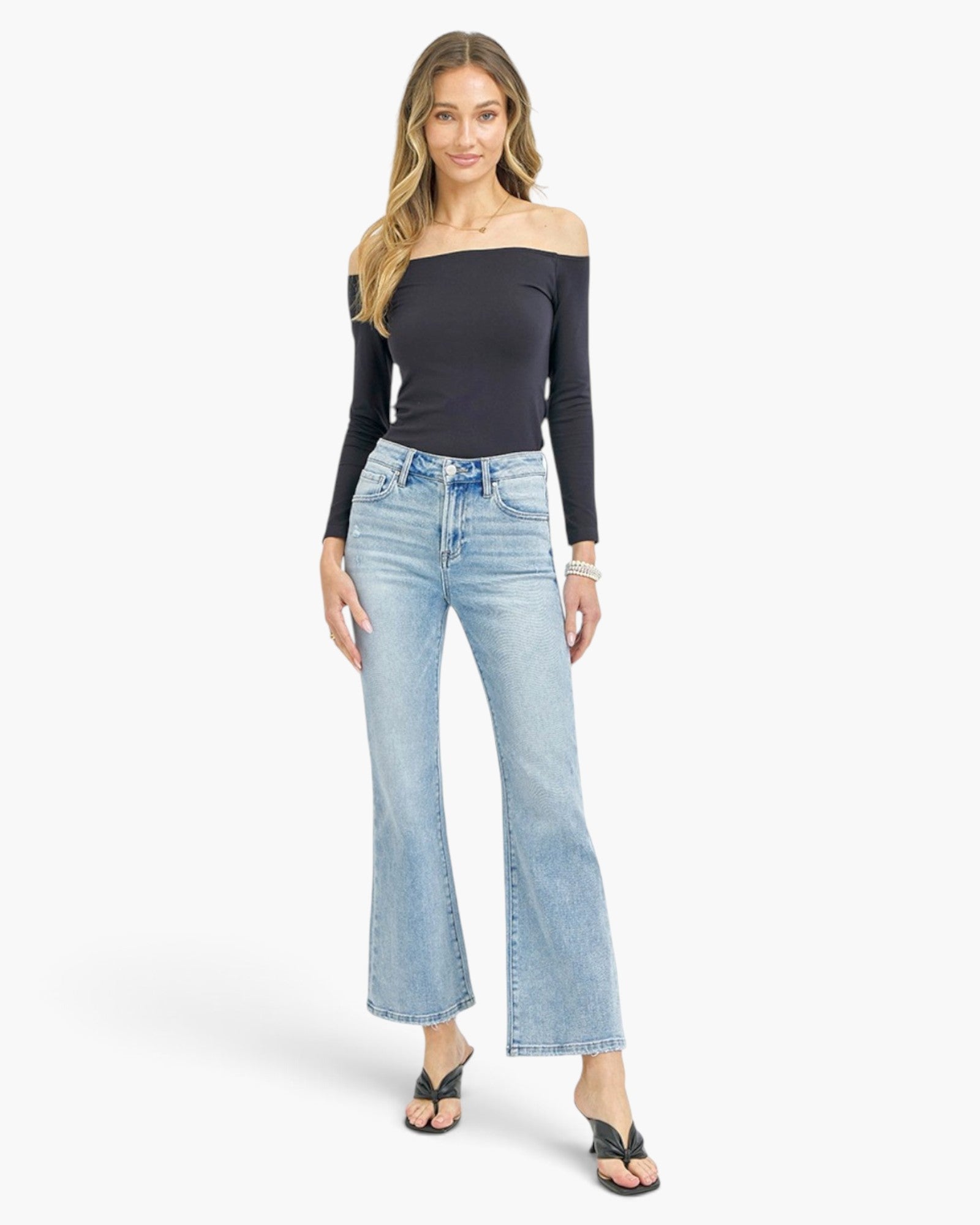 A woman modeling a black off-the-shoulder long-sleeve top and light-wash blue flare jeans with black sandals, photographed against a plain light gray studio background.