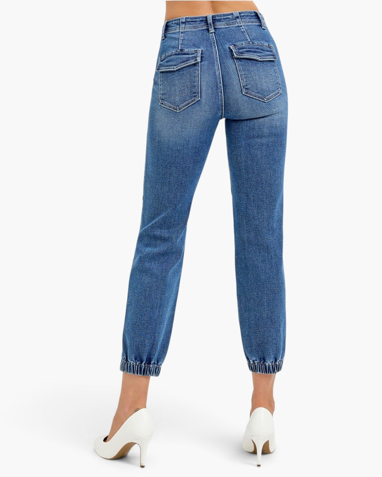 Rear view of a model wearing medium-wash blue cropped jeans with cuffed hems and white pointed-toe heels against a light gray studio background.