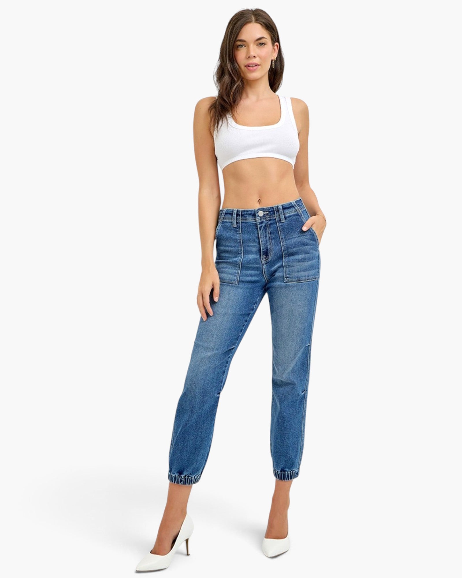 A model wearing a pair of high-waisted, straight-leg blue jeans with a cropped hem, styled with a white crop top and white pointed-toe heels against a light gray studio background.